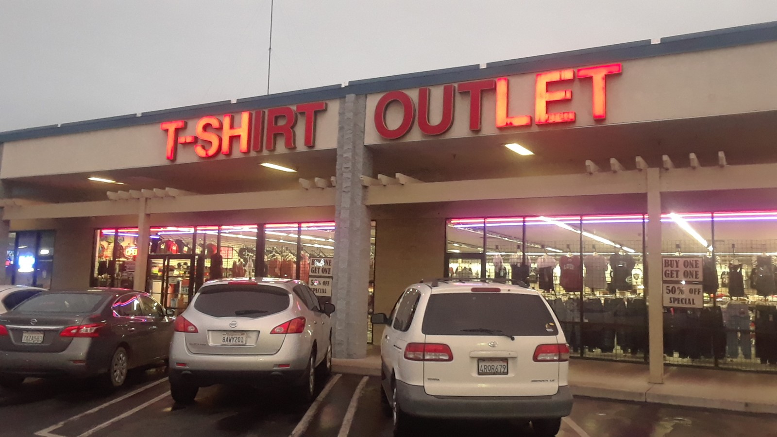 Front of T-Shirt Outlet at dusk with the red sign lit